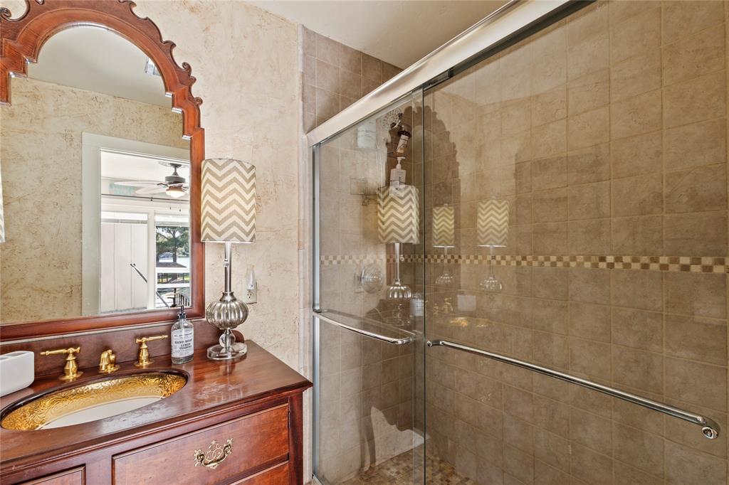 Bathroom featuring a wood vanity with a recessed basin, gold-tone faucet hardware, arched wall mirror, and a walk-in shower enclosure with tile surround