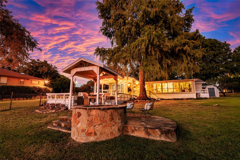 Expansive grounds featuring a stone-clad fire pit, a covered outdoor pavilion with a raised wooden deck, and mature shade trees