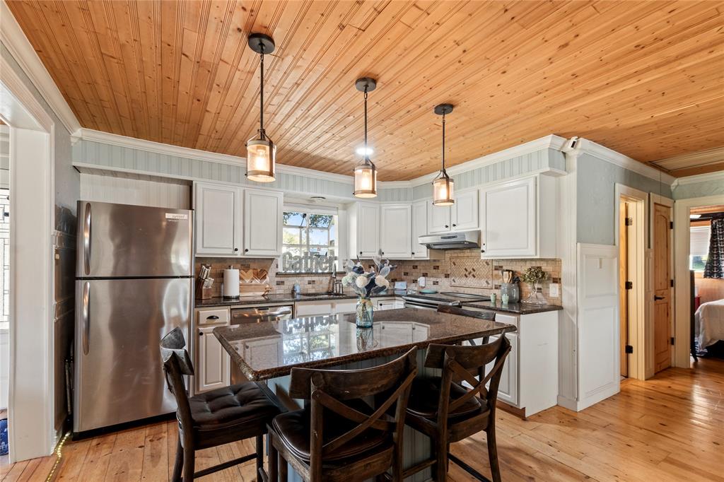 Kitchen featuring natural wood-plank ceiling, white cabinetry, central island with stone countertop, stainless steel refrigerator, and three pendant light fixtures Kitchen featuring natural wood-plank ceiling, white cabinetry, central island with stone countertop, stainless steel refrigerator, and three pendant light fixtures
