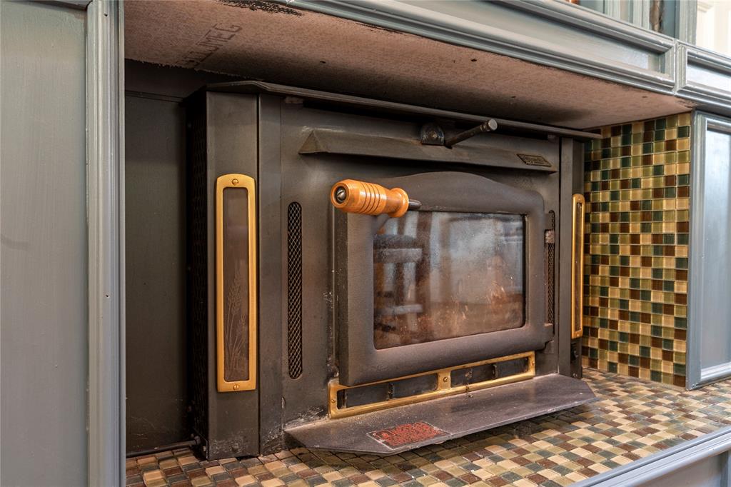 Wood-burning stove insert featuring a matte black finish, brass accents, a glass viewing panel, and a wood-toned handle, framed by mosaic tile surfacing and custom cabinetry Wood-burning stove insert featuring a matte black finish, brass accents, a glass viewing panel, and a wood-toned handle, framed by mosaic tile surfacing and custom cabinetry