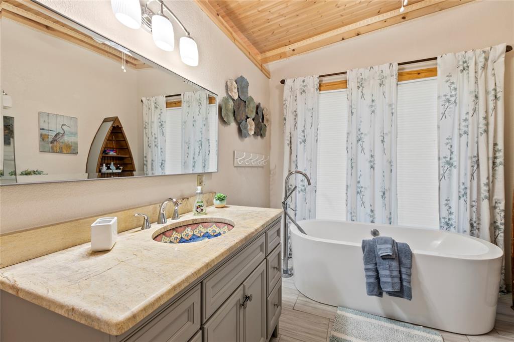Freestanding soaking tub with a floor-mounted faucet, stone countertop with an integrated patterned basin, grey cabinetry, and wood-paneled ceiling