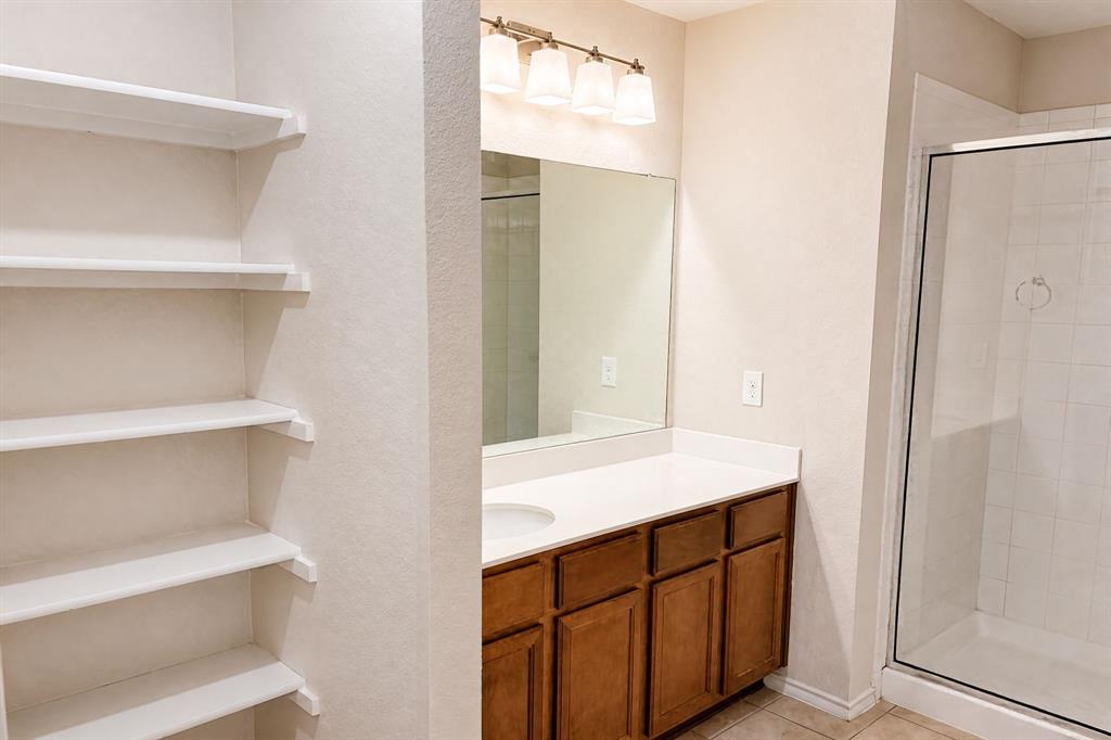 Bathroom featuring stained wood cabinetry, white countertop, integrated shelving, four-light vanity fixture, and a glass-enclosed shower with tiled surround