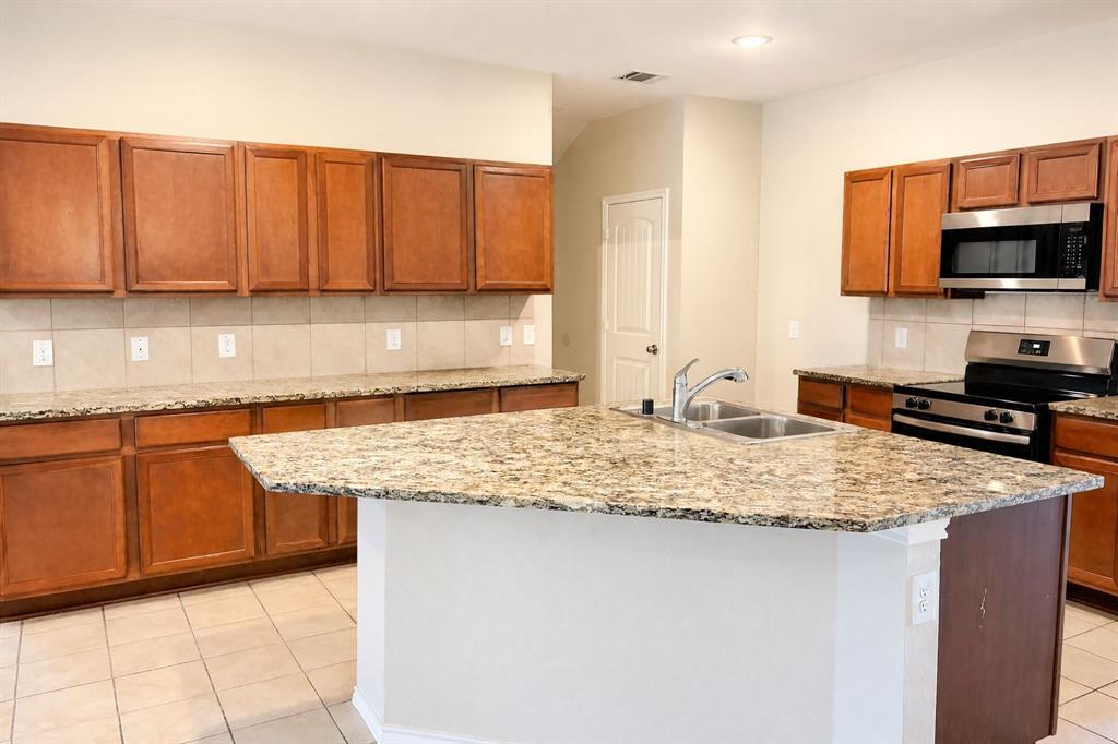 Kitchen featuring natural wood cabinetry, stone-patterned countertops, a central island with a double-basin sink, tile backsplash, and stainless steel appliances