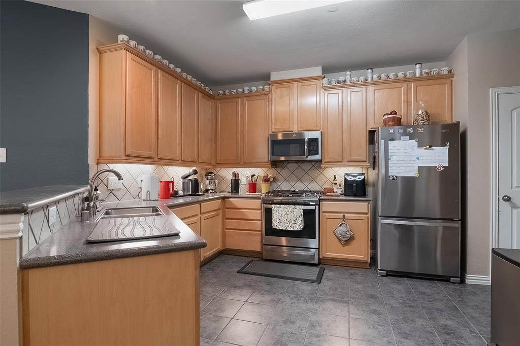 Kitchen featuring natural wood cabinetry, stainless steel appliances, a diagonal tile backsplash, dark solid-surface countertops, and tile flooring