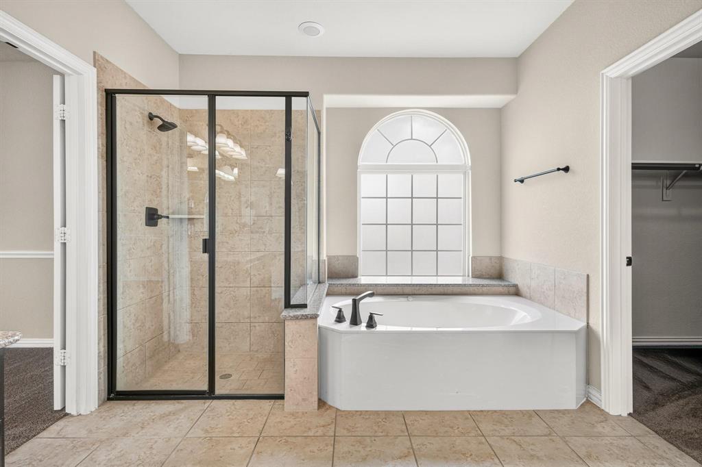 Glass-enclosed shower with tile surround, soaking tub with arched window, and stone-finish tile flooring