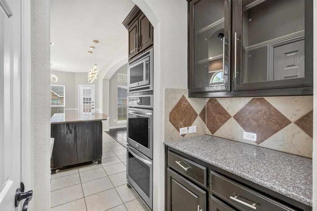 Kitchen featuring dark wood cabinetry, built-in stainless steel double ovens and microwave, speckled stone-finish countertops, decorative tile backsplash, and ceramic tile flooring