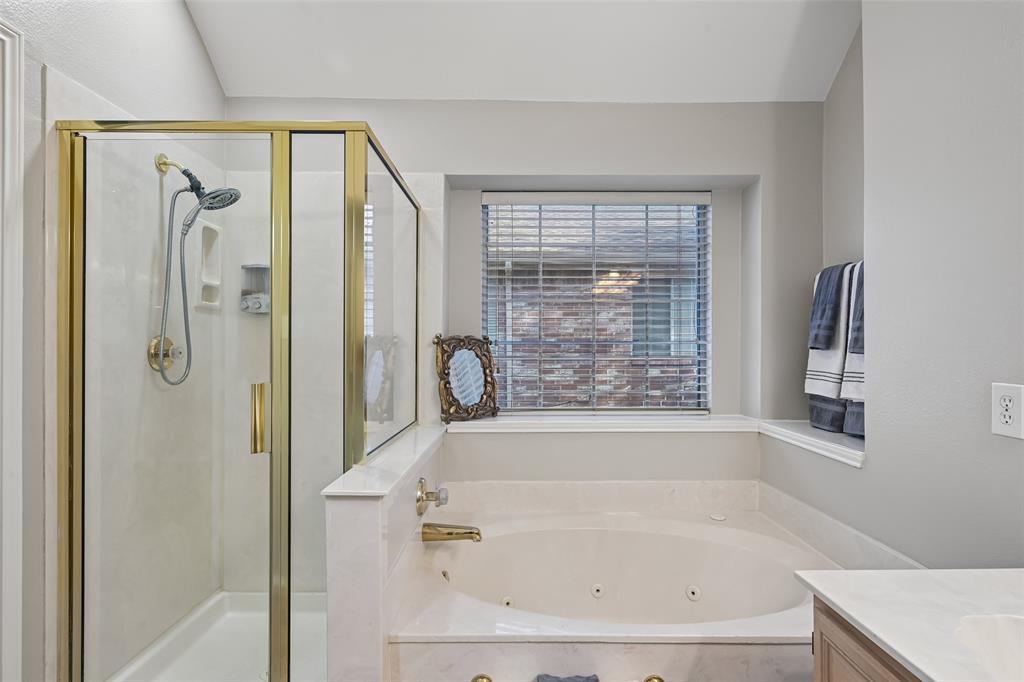 Jetted soaking tub with stone-finish surround, glass-enclosed shower with gold-tone frame, handheld shower fixture, and recessed window sill