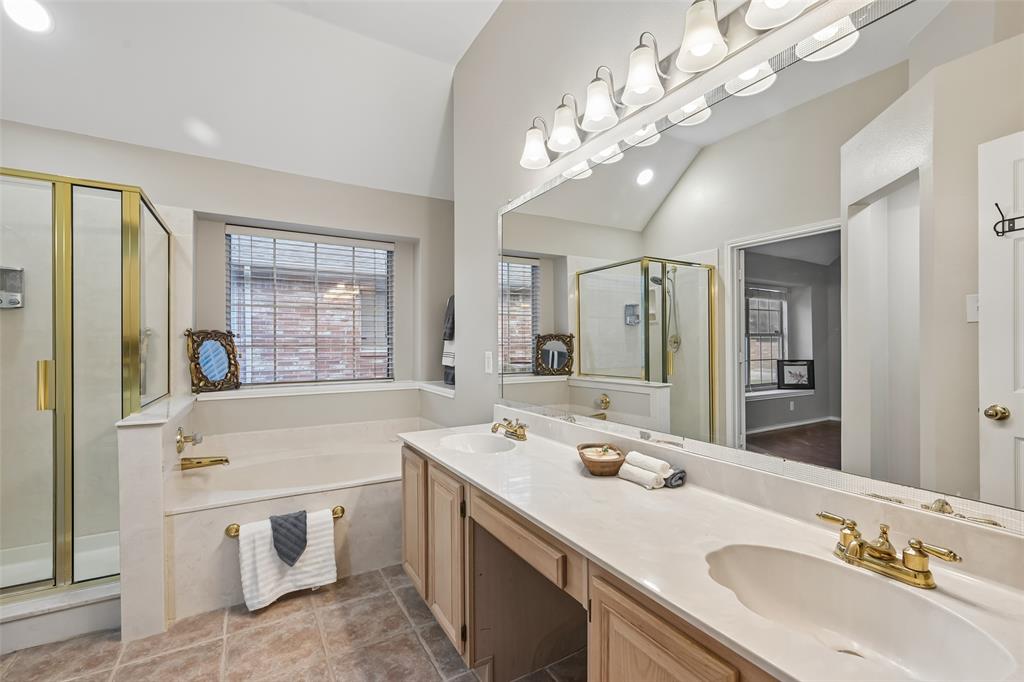 Bathroom featuring dual-sink vanity with light-tone wood cabinetry, solid surface countertop, brass-finish fixtures, framed glass-enclosed shower, and built-in soaking tub