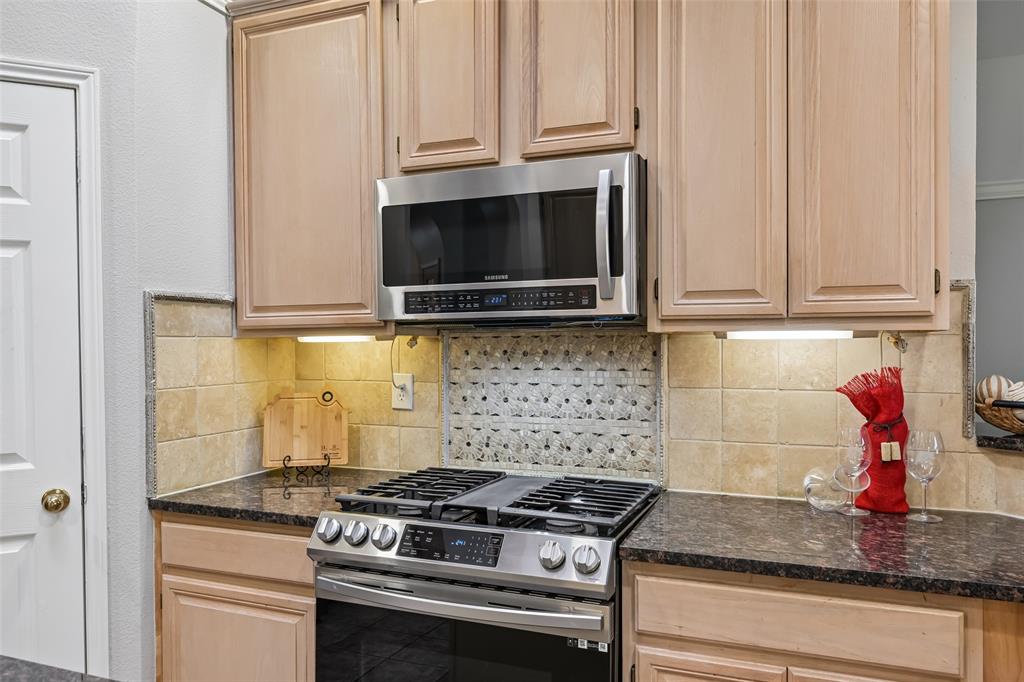Light-toned wood cabinetry, stainless steel gas range, stainless steel microwave, stone-finish countertops, and decorative tile backsplash