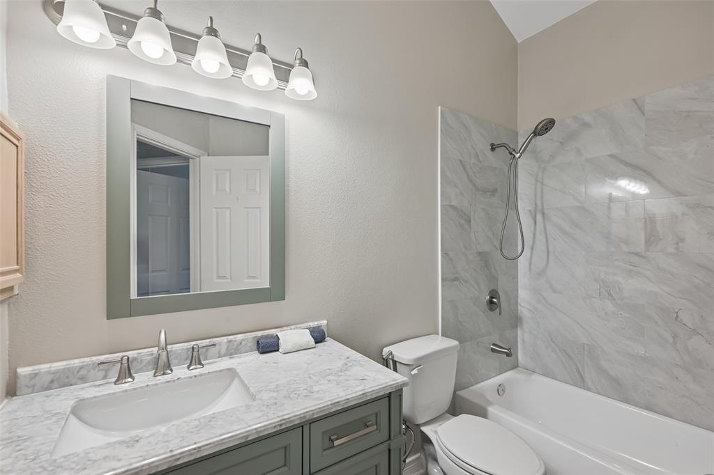 Bathroom vanity featuring a stone-look countertop with an integrated rectangular sink, five-light fixture, framed mirror, and tiled shower surround