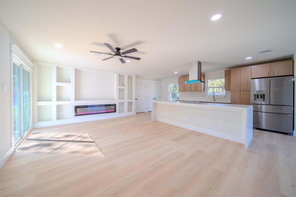 Open-concept interior featuring light wood-finish flooring, built-in shelving, an electric wall-mounted fireplace, a kitchen island with stone-style countertops, and stainless steel appliances