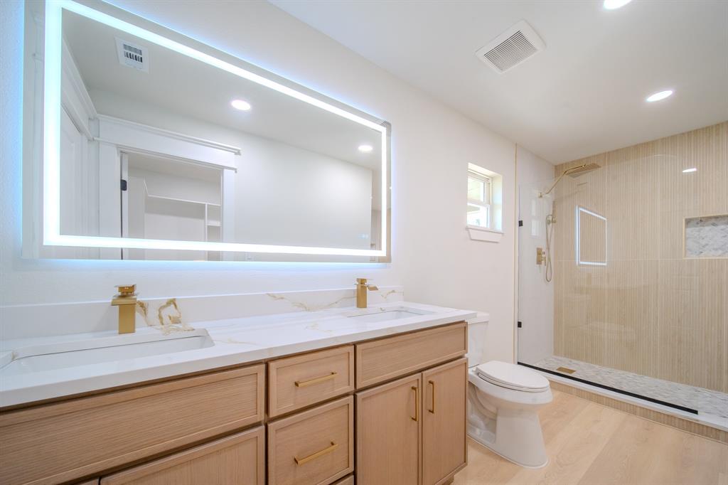 Dual vanity featuring light wood cabinetry, white stone-finish countertop, gold-tone hardware, large integrated LED mirror, and a glass-enclosed walk-in shower