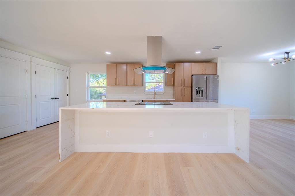 Kitchen featuring a waterfall-edge stone island, wood-finish cabinetry, stainless steel range hood, built-in refrigerator, and wood-finish flooring