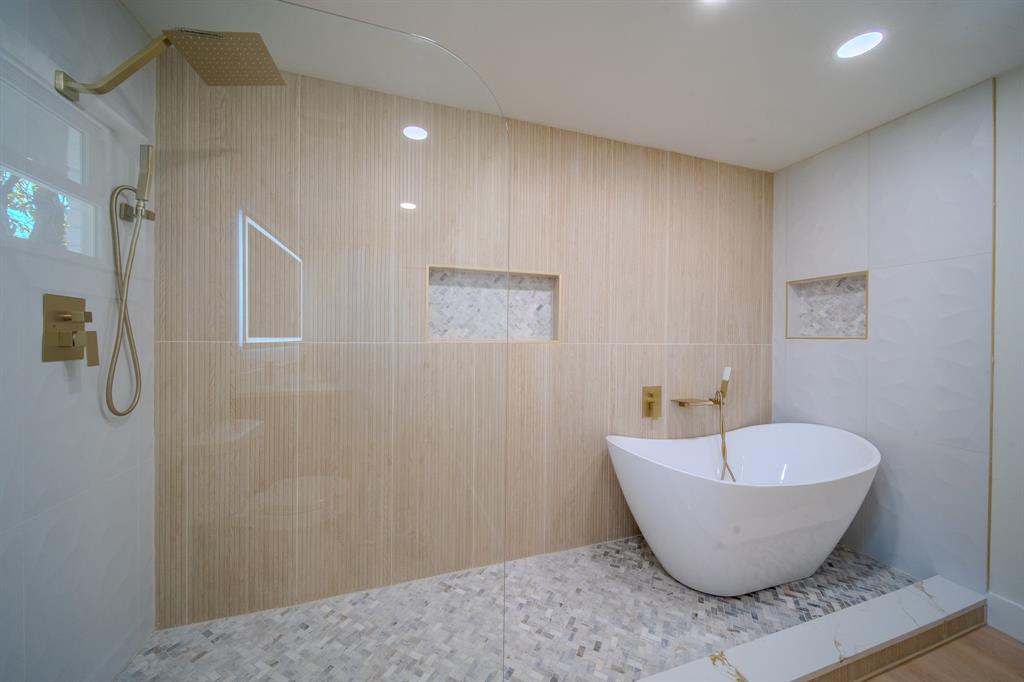 Freestanding soaking tub featuring gold-tone hardware, large-format textured wall tile, glass-enclosed shower area with rain-style showerhead, recessed wall niches, and intricate herringbone-patterned floor tile