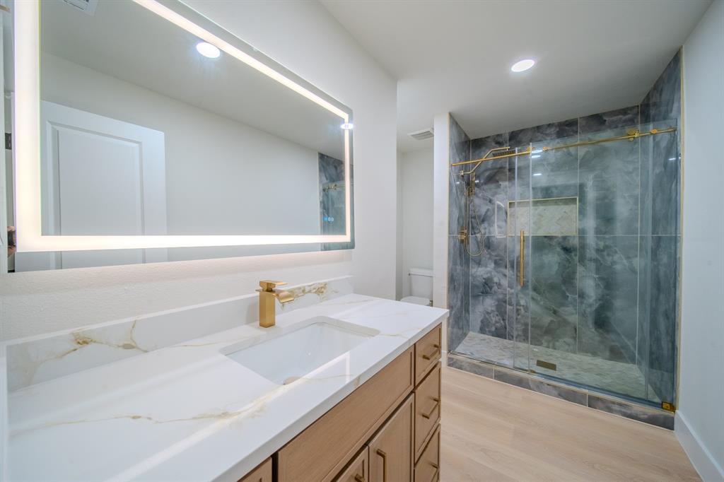 Modern vanity with gold-tone hardware and quartz countertop featuring a rectangular under-mount sink, integrated LED mirror, and walk-in shower with stone-tile surround and gold-tone fixtures