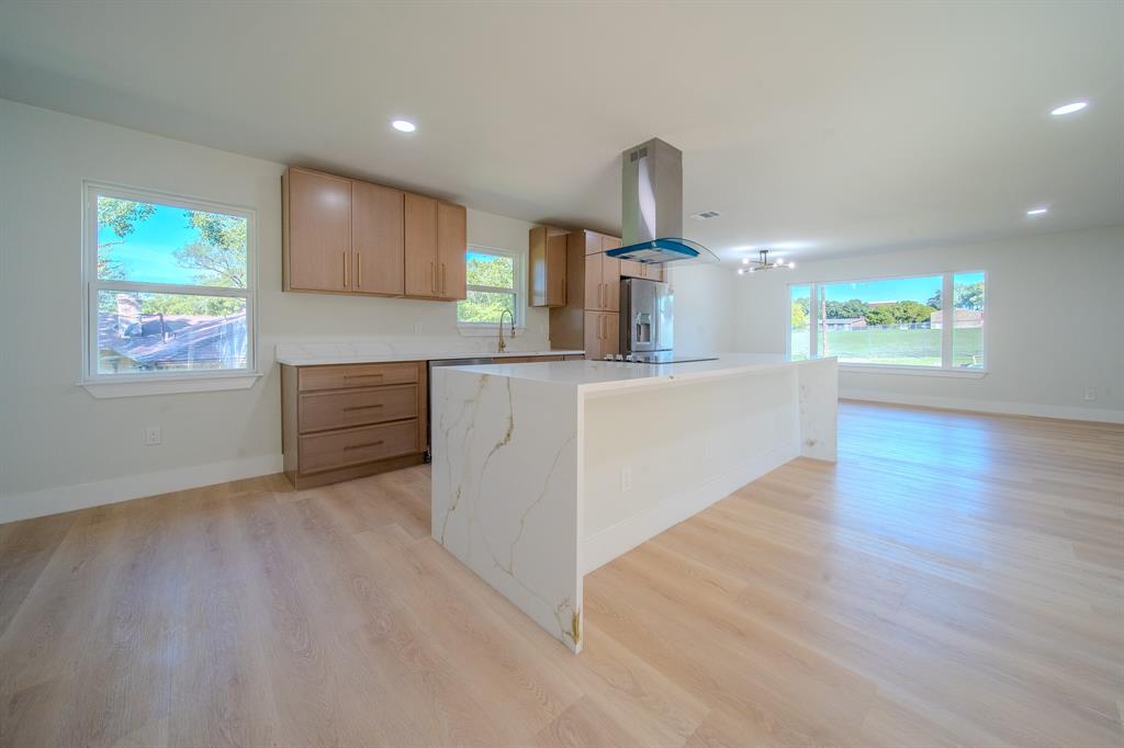 Kitchen featuring wood-finish flooring, a waterfall-edge island with stone-finish countertops, stainless steel chimney-style range hood, and flat-panel cabinetry