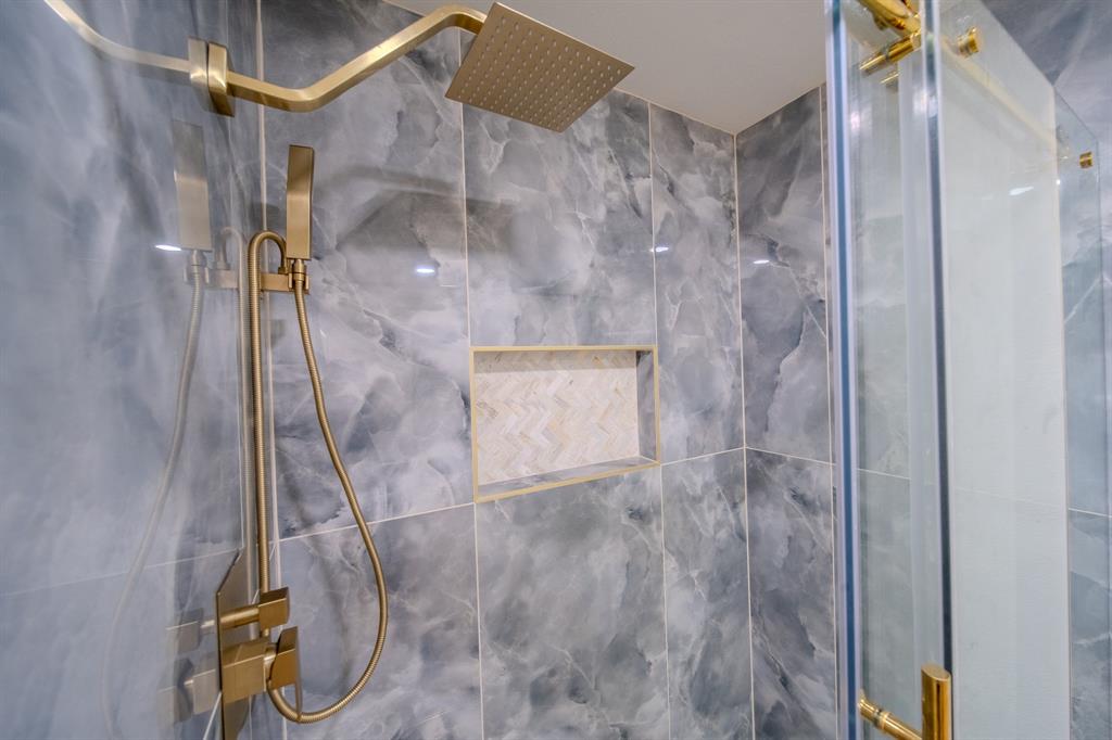 Walk-in shower featuring large-format grey stone-patterned wall tile, brass-finish rainfall and handheld shower fixtures, a built-in recessed niche with chevron-pattern inlay, and a glass enclosure