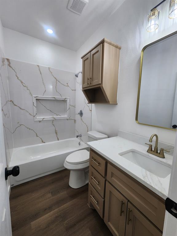 Bathroom featuring wood-finish flooring, a vanity with a stone-top integrated sink and gold-tone faucet, a matching wall-mounted cabinet, and a tiled shower-tub surround with an inset niche