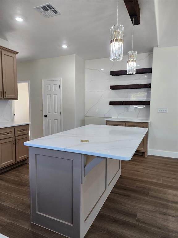 Kitchen featuring a central island with a solid surface countertop, wood-finish flooring, custom shaker-style cabinetry, floating shelves, and crystal pendant lighting Kitchen featuring a central island with a solid surface countertop, wood-finish flooring, custom shaker-style cabinetry, floating shelves, and crystal pendant lighting