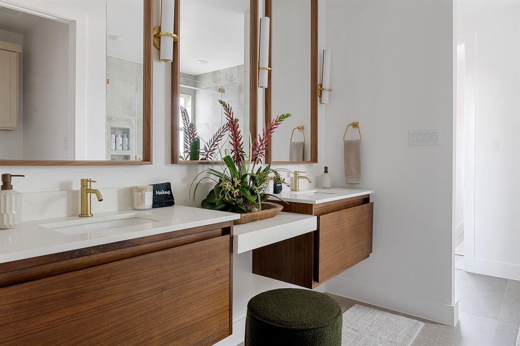 Floating dual-vanity with wood cabinetry, white stone countertops, integrated sinks, brass-toned fixtures, and vertical-frame mirrors with matching sconces
