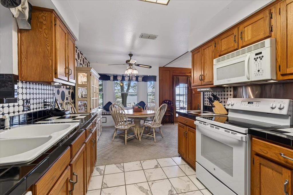 Kitchen featuring wood cabinetry, white appliance suite, geometric patterned tile backsplash, dark countertops, and integrated tile flooring