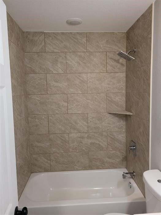 Bathtub and shower combination featuring large-format neutral-toned tile surround, built-in corner shelving, chrome rainfall showerhead, and matching chrome faucet fixture
