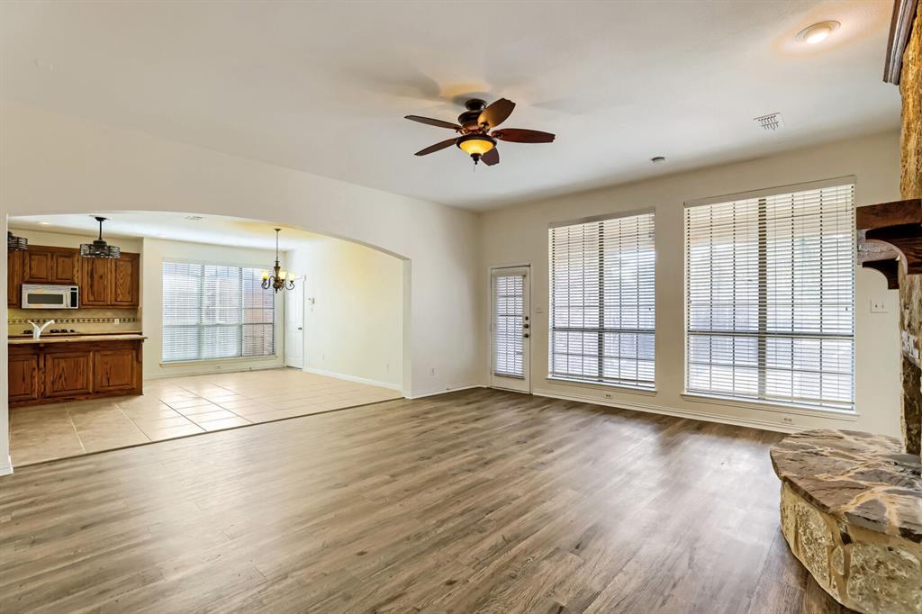 Open-concept interior featuring wood-finish flooring, a stone-accented fireplace hearth, large windows with horizontal blinds, a vaulted ceiling with a central fan, and ceramic tile flooring in the kitchen area