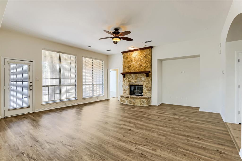Living area featuring wood-finish flooring, floor-to-ceiling stone fireplace with a timber mantel, high ceilings, ceiling fan, and large windows with integrated blinds