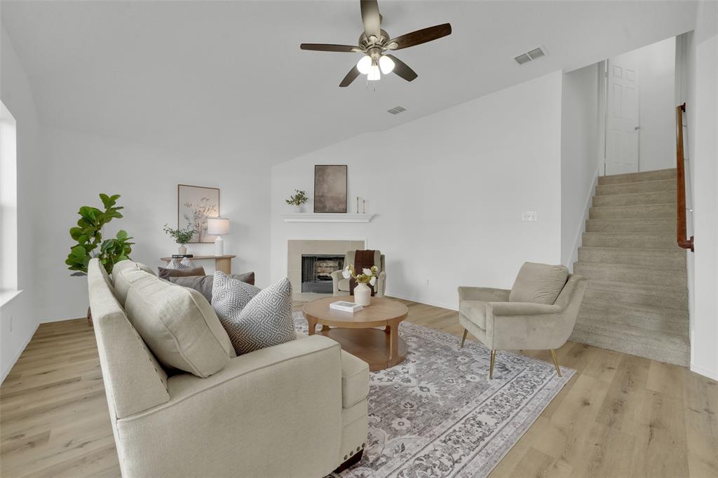 Living area featuring wood-finish flooring, vaulted ceilings, a central fireplace with a neutral-toned surround, a ceiling fan, and carpeted staircase
