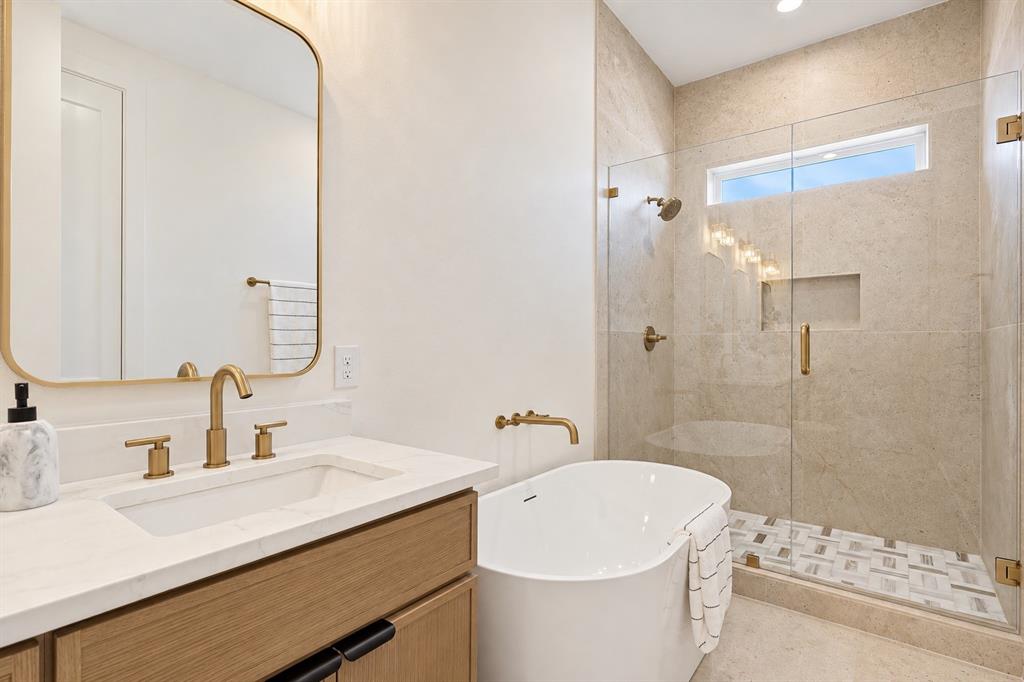 Freestanding tub with wall-mounted faucet, glass-enclosed shower with contrasting tile floor, single vanity with wood-finish cabinetry, and gold-tone fixtures throughout