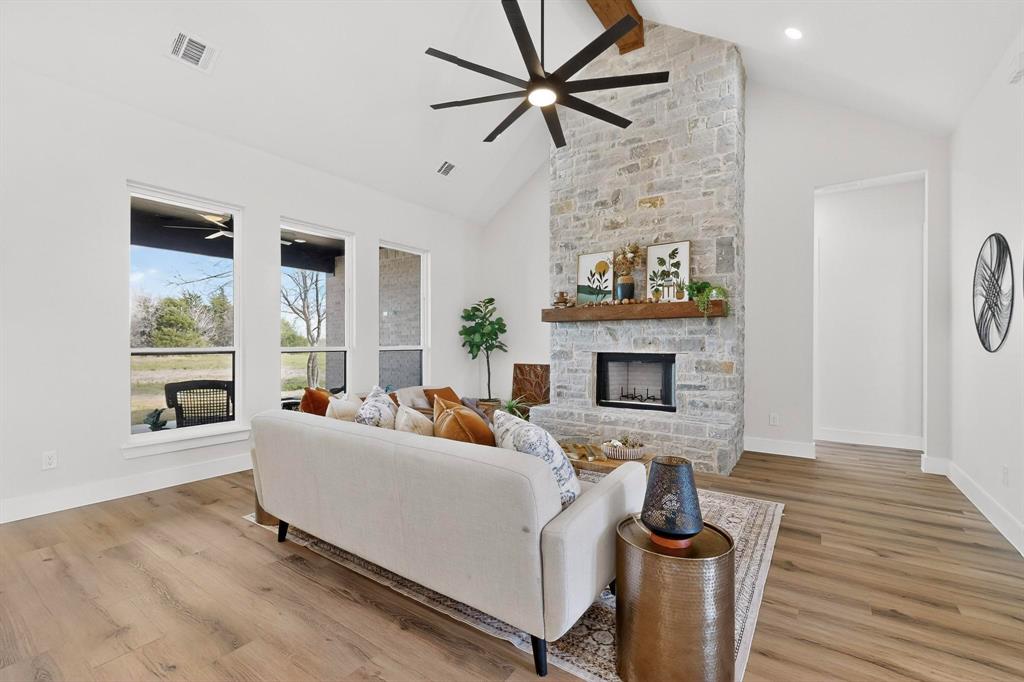 Vaulted ceiling with an exposed wood beam, floor-to-ceiling stone fireplace, prominent wood-finish flooring, multi-pane floor-to-ceiling windows, and a large-format multi-blade ceiling fan