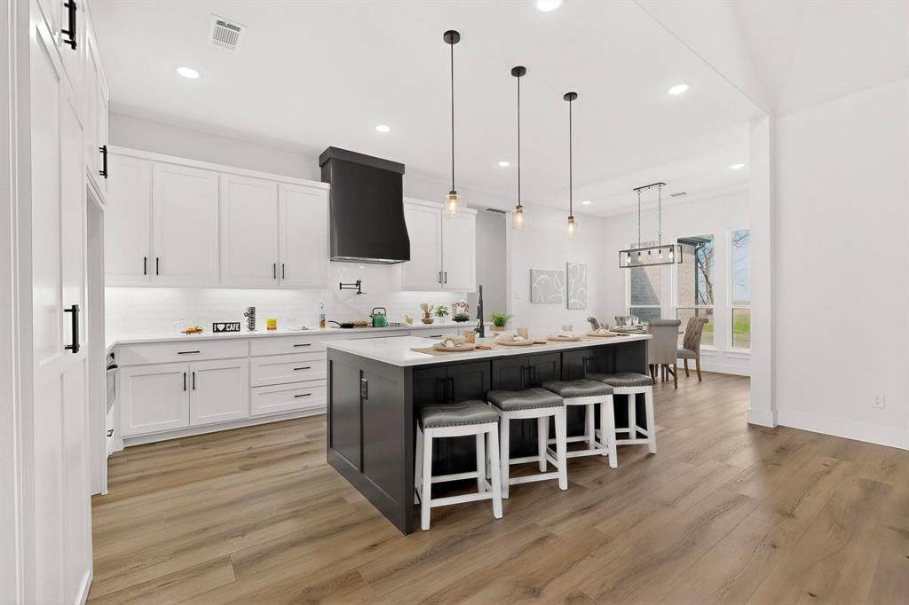 Kitchen featuring wood-finish flooring, white cabinetry, a center island with a contrasting dark base, a built-in range hood, and three pendant lights