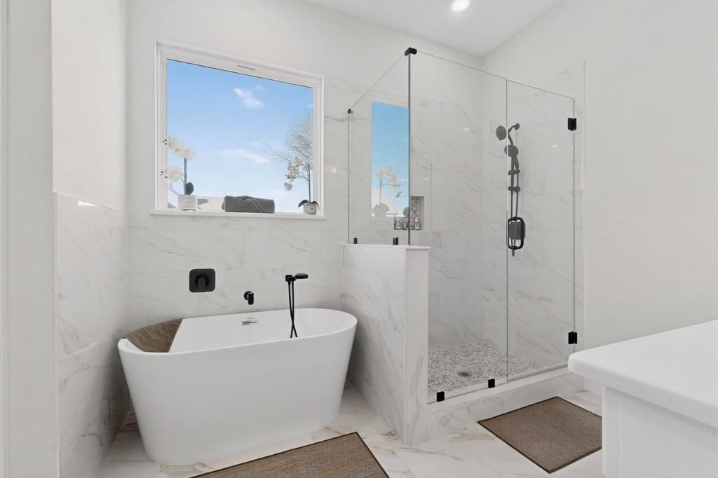 Freestanding soaking tub with matte black wall-mounted faucet, walk-in glass-enclosed shower with pebble-tile flooring, large-format tile wall surround, and recessed shower niche