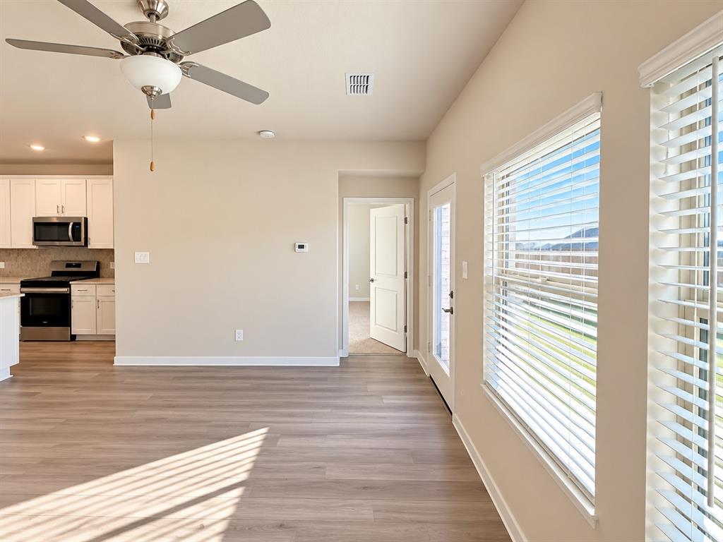 Wood-finish flooring, ceiling fan with integrated light, windows with horizontal blinds, and kitchen featuring white cabinetry and stainless steel appliances