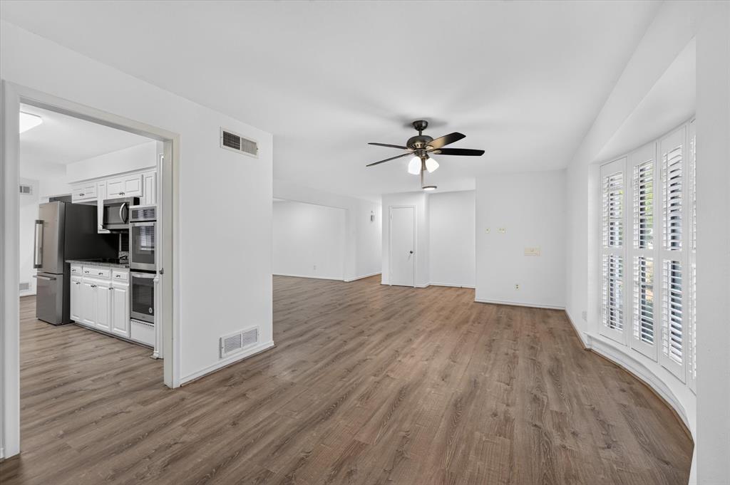Open-concept interior featuring wood-finish flooring, a bay window with louvered shutters, and a ceiling fan with integrated lighting Open-concept interior featuring wood-finish flooring, a bay window with louvered shutters, and a ceiling fan with integrated lighting