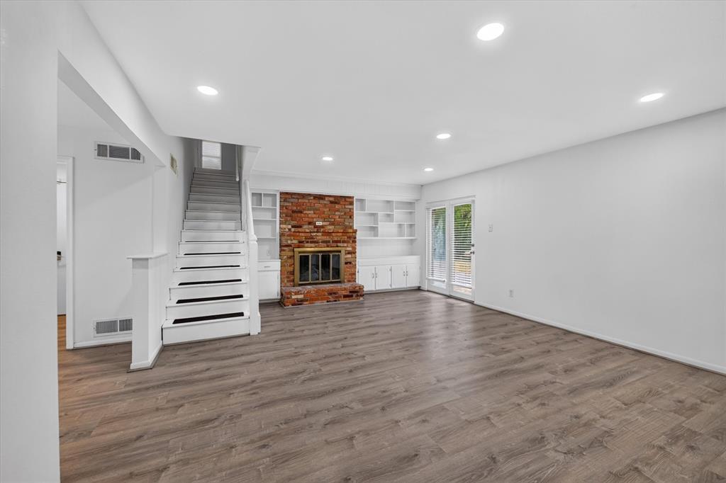 Open living area featuring wood-finish flooring, recessed lighting, red brick fireplace, built-in cabinetry, and double glass doors Open living area featuring wood-finish flooring, recessed lighting, red brick fireplace, built-in cabinetry, and double glass doors