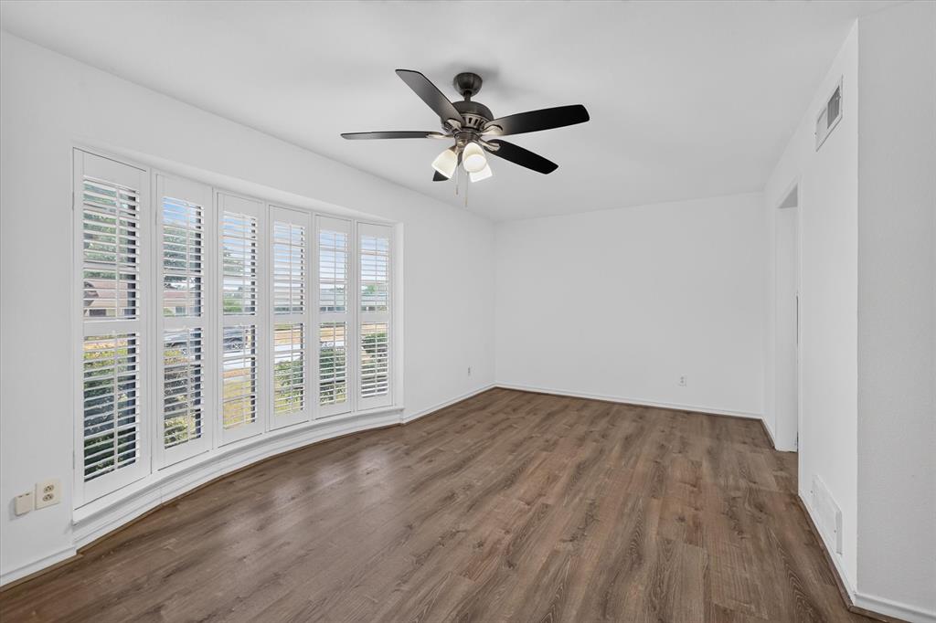 Room featuring wood-finish flooring, a bay window with white plantation shutters, and a central ceiling fan with integrated lighting Room featuring wood-finish flooring, a bay window with white plantation shutters, and a central ceiling fan with integrated lighting