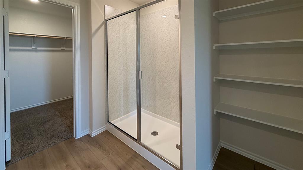Walk-in shower with clear glass enclosure and textured wall panels, wood-finish flooring, built-in shelving, and an additional walk-in closet with carpeted flooring and hanging rods