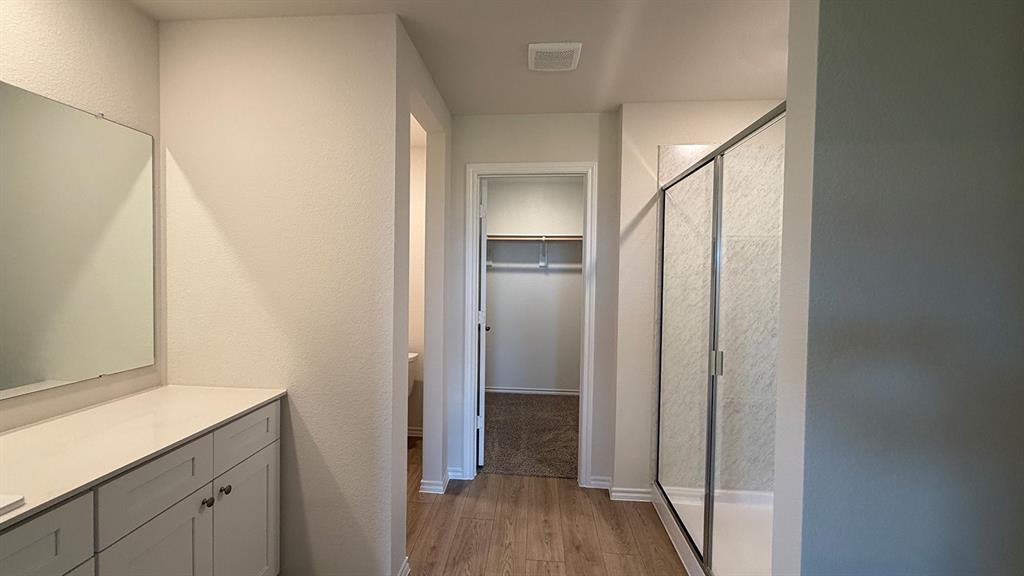 Light-filled bathroom featuring a glass-enclosed shower, a vanity with light-toned cabinetry, a spacious countertop, wood-finish flooring, and an open closet with shelving