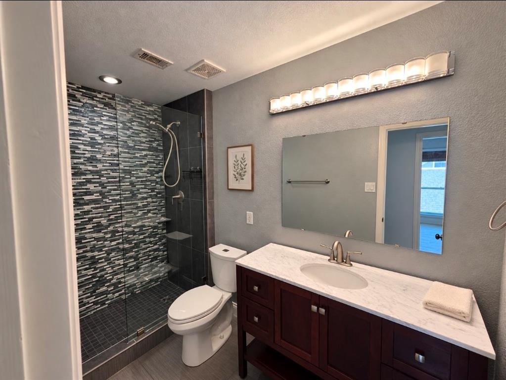 Bathroom featuring a walk-in shower with a multi-textured stone-tile surround, dark wood-finish vanity with a stone countertop, oval undermount sink, and full-width wall-mounted lighting