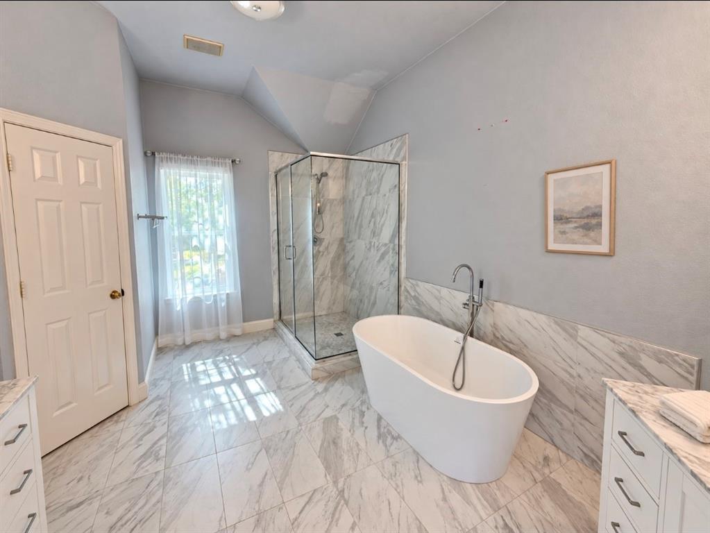 Freestanding soaking tub with a floor-mounted faucet, glass-enclosed shower, stone-finish tile flooring, and stone-finish wall surrounds