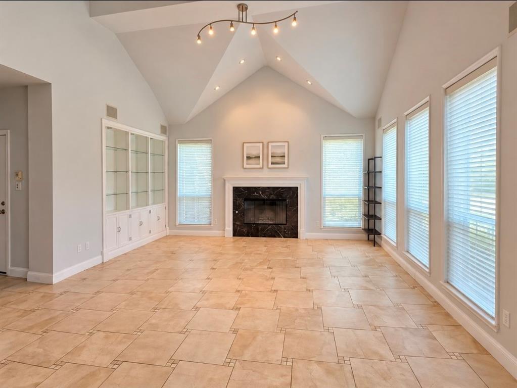 Vaulted ceilings, stone-surround fireplace with mantel, built-in display cabinetry with glass shelving, tile flooring, and multiple tall windows with blinds Vaulted ceilings, stone-surround fireplace with mantel, built-in display cabinetry with glass shelving, tile flooring, and multiple tall windows with blinds