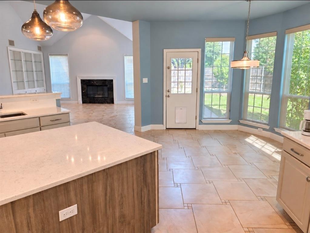 Kitchen area featuring a center island with light-toned stone countertop, ceramic tile flooring, built-in display cabinetry, vaulted ceiling, and fireplace with a dark stone surround Kitchen area featuring a center island with light-toned stone countertop, ceramic tile flooring, built-in display cabinetry, vaulted ceiling, and fireplace with a dark stone surround