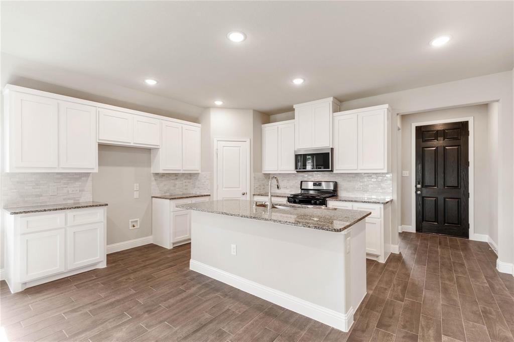 Kitchen featuring white shaker-style cabinetry, stone countertops, a center island with a stainless steel sink, wood-finish flooring, and a tiled backsplash