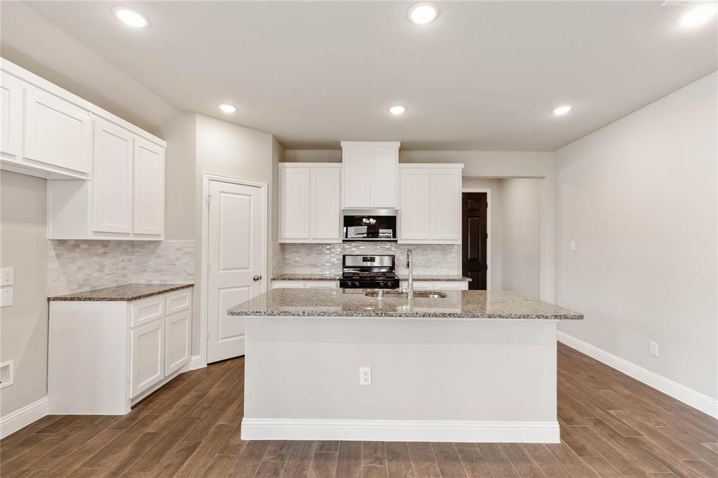 Kitchen featuring white shaker-style cabinetry, stone-surface countertops, a tiled backsplash, wood-finish flooring, and a central island with an integrated stainless steel sink