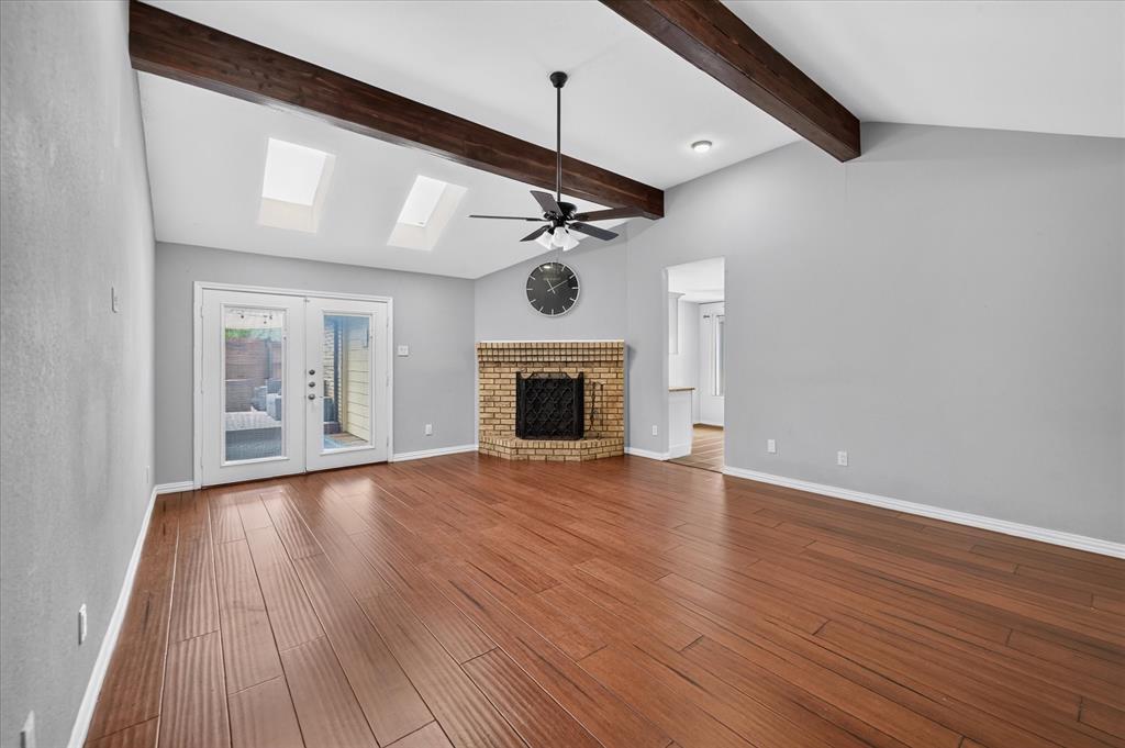 Vaulted ceiling with exposed wood beams, wood-finish flooring, brick corner fireplace, dual skylights, and double-glass paneled exterior doors