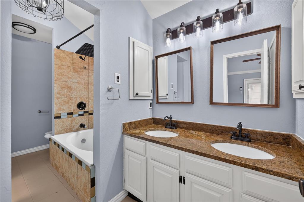Dual-sink vanity with stone countertop and matte black fixtures, custom wall-mounted mirrors, and soaking tub featuring stone-tile surround