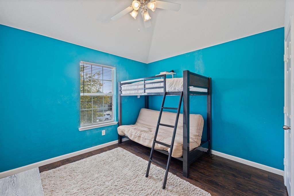 A vibrant bedroom featuring striking turquoise accent walls, a vaulted ceiling, and rich wood-look flooring. A window with blinds brings in natural light while maintaining privacy, and the contrasting white trim adds a crisp, clean finish. The distinctive wall color creates a lively atmosphere, making this room an inviting space with plenty of personality.