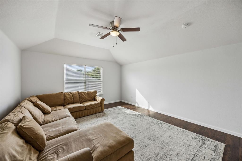 A spacious upstairs living area featuring a vaulted ceiling, ceiling fan, and rich wood-look flooring that extends throughout the room. Neutral wall colors and a large window invite natural light inside, creating a bright, open atmosphere. The generous floor space offers flexibility for a secondary living area, game room, media room, or comfortable retreat.