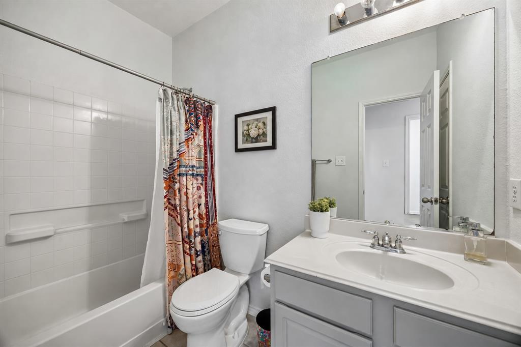 This full bathroom on the first floor features a tub and shower combination with white tile surround, a single-sink vanity, and a large wall mirror with bright vanity lighting. Gray cabinetry complements the light countertop and neutral wall color, while tile flooring provides a durable, easy-maintenance finish. The functional layout offers everyday convenience and a clean, timeless look.